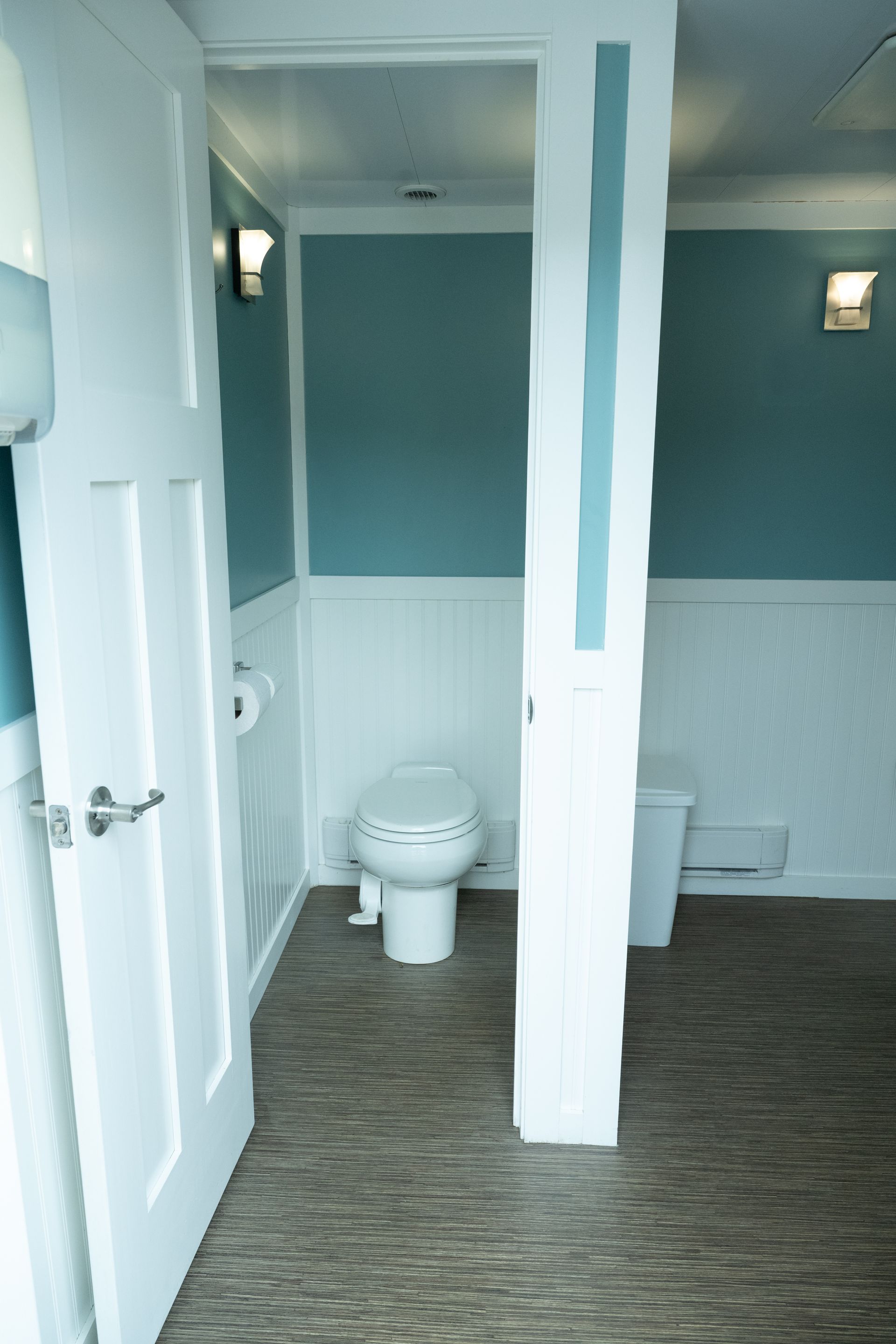 Interior of a portable restroom with a toilet, paper, and waste bin. Blue and white decor. Door open.