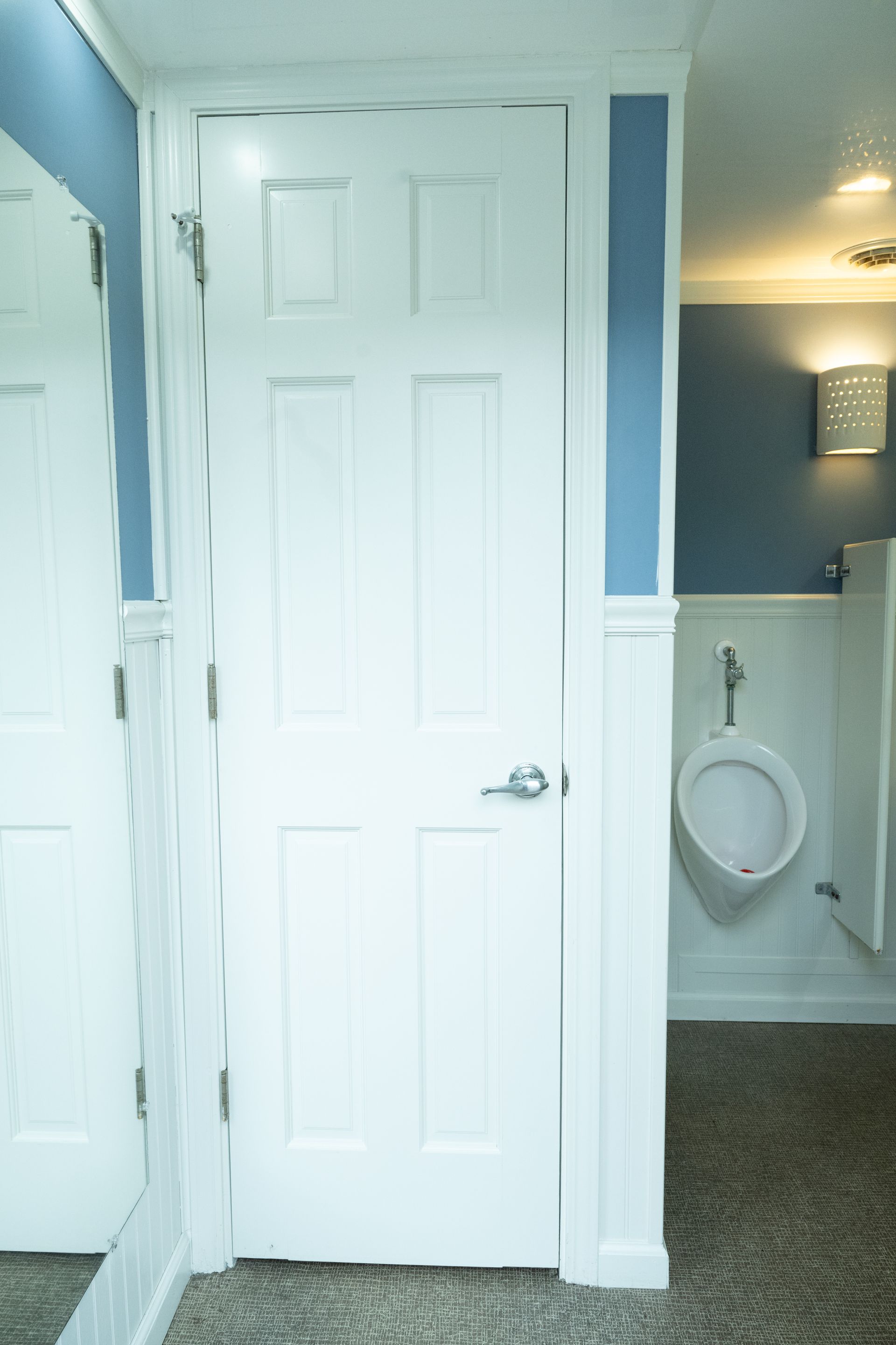 White door in a public restroom with a urinal visible. Blue walls, and a mirror are present.