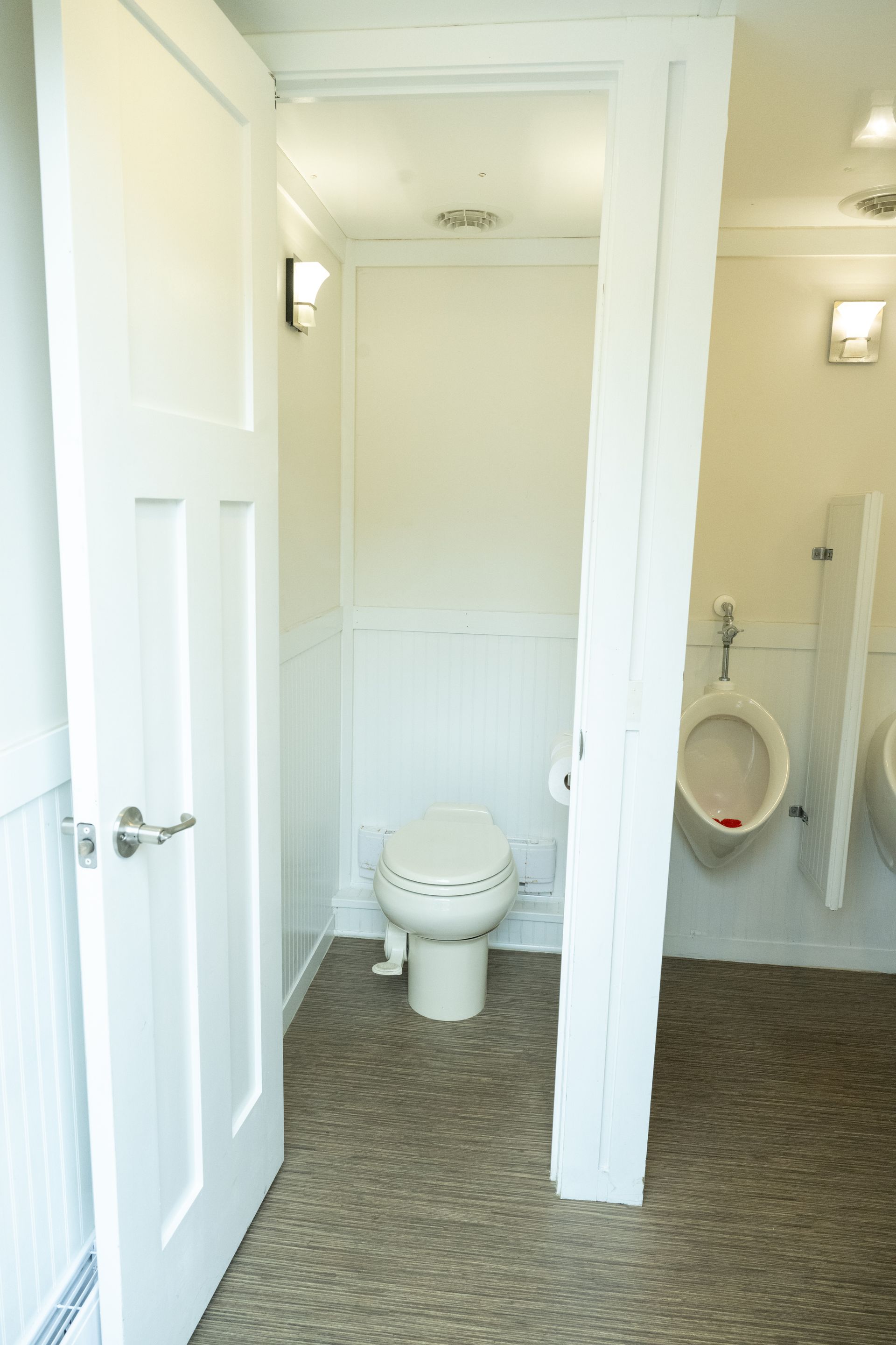 Interior of a white portable restroom, showing a toilet and urinals. The floor is speckled brown.