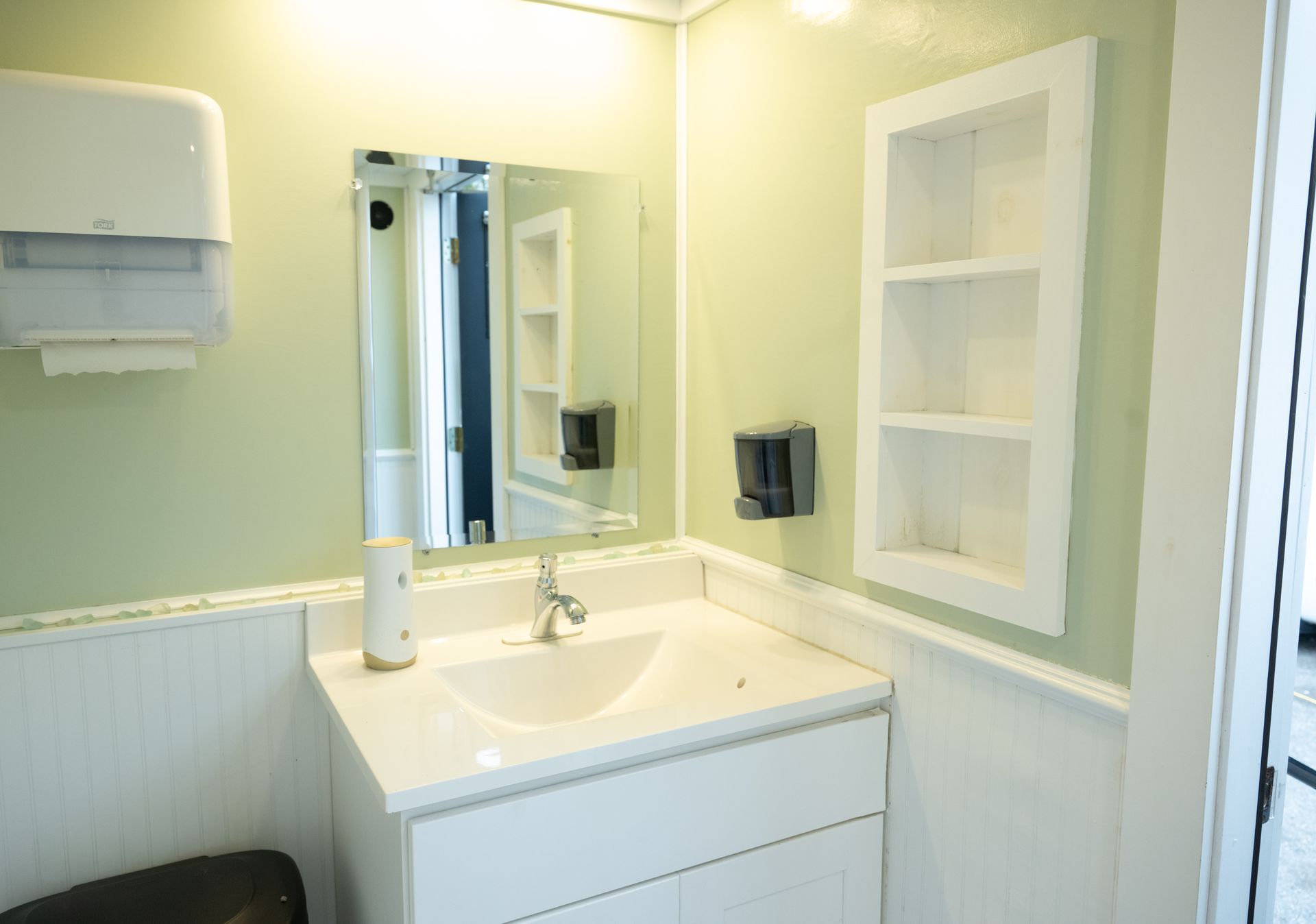 Bathroom interior with sink, mirror, and shelves. Light green walls, white cabinets, and wooden paneling.
