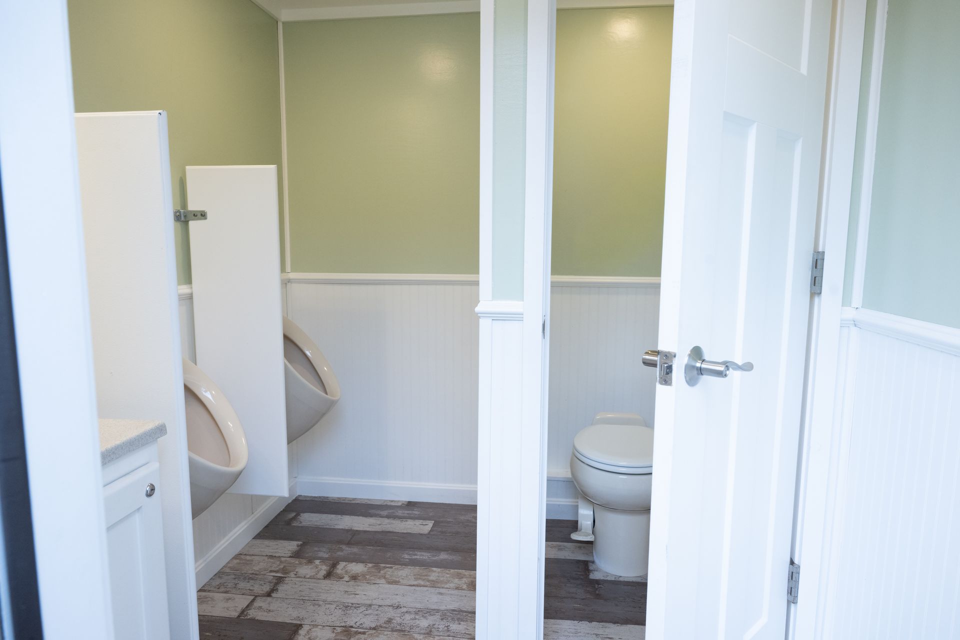 Public restroom with a urinal and toilet, featuring white and sage green walls, and wood-look flooring.