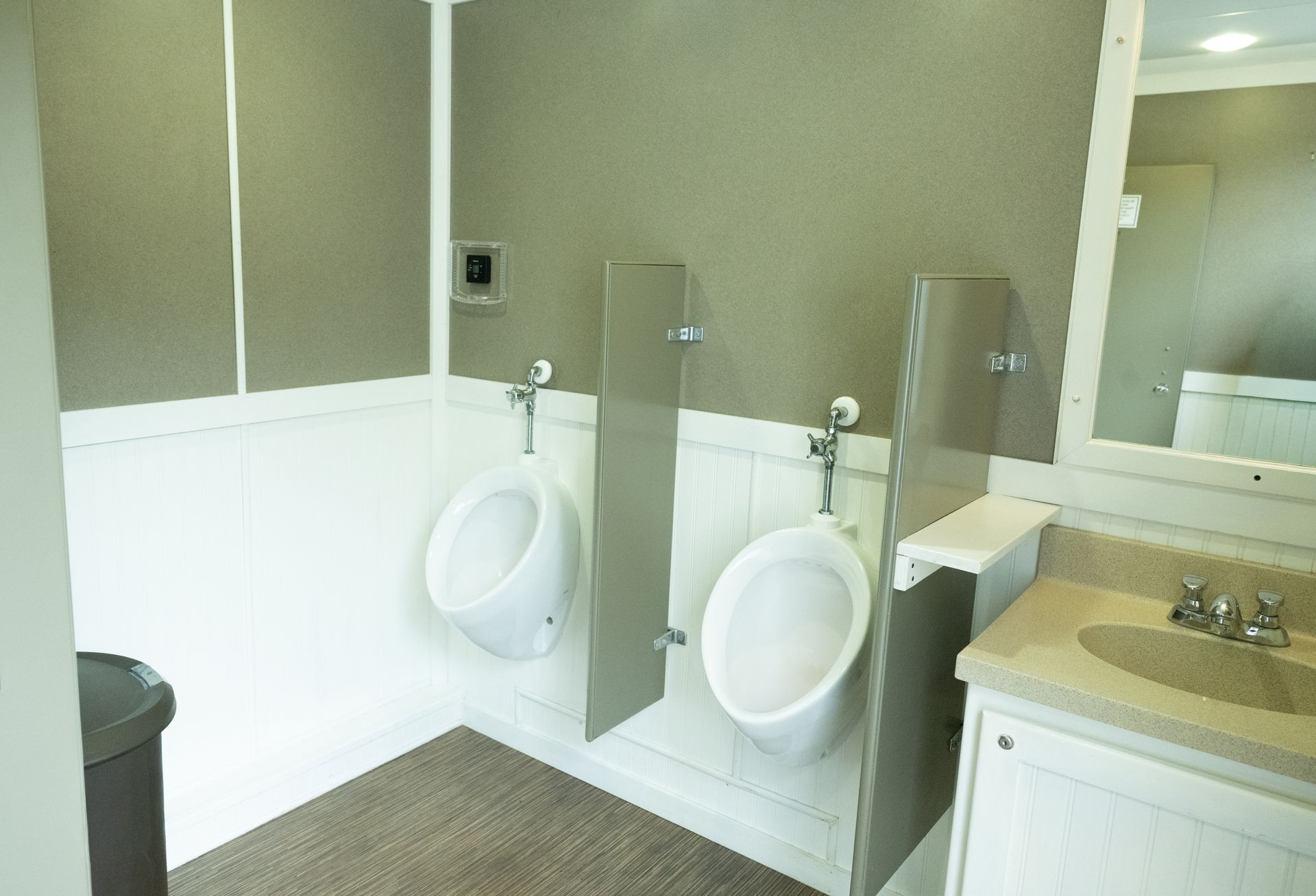 Two urinals in a public restroom with grey dividers, beige countertops, and a mirror.