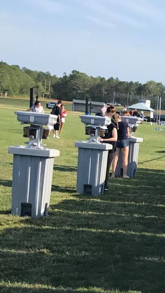 People stand at a row of portable handwashing stations on a grassy field on a sunny day.