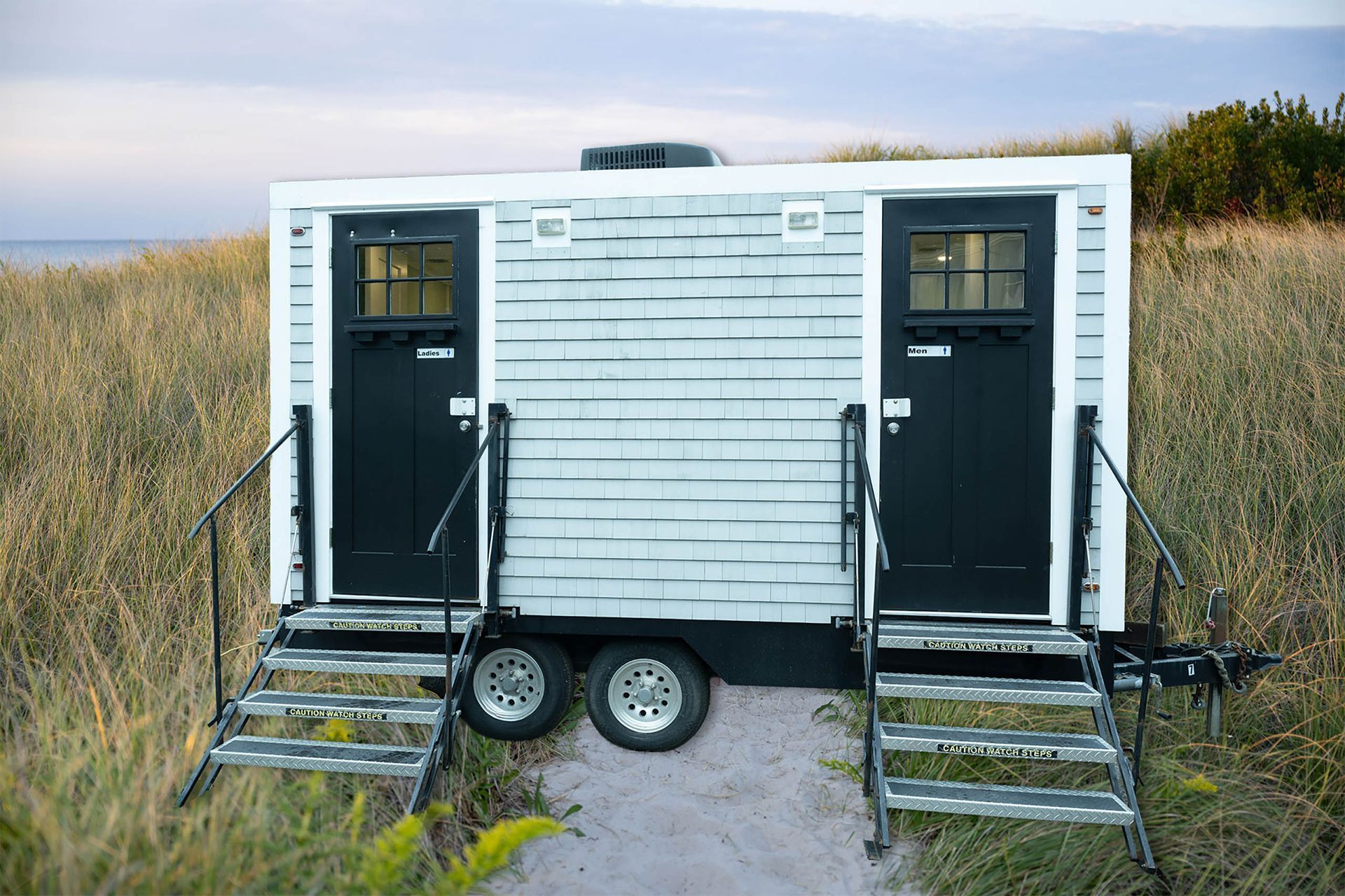 White portable cabin with two black doors and stairs, set on a sandy path in tall grass near the water.