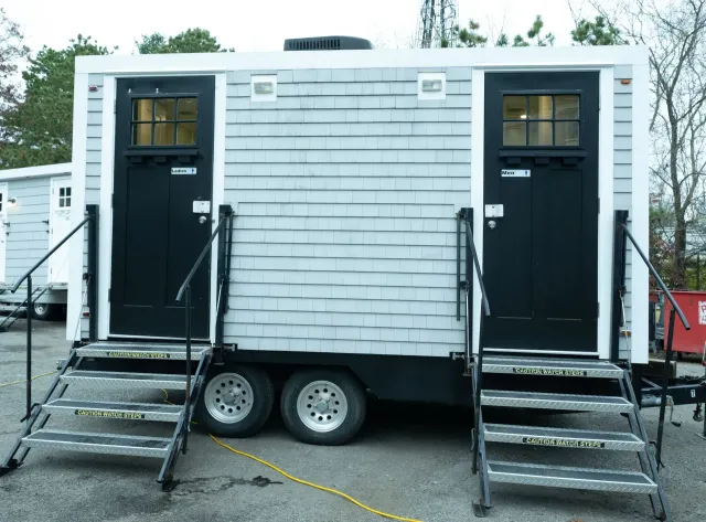 Portable restroom trailer with light gray shingled siding, two black doors with windows, and metal entry stairs.