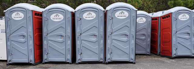 A row of portable toilets in red and gray colors sits on a gravel surface outdoors.