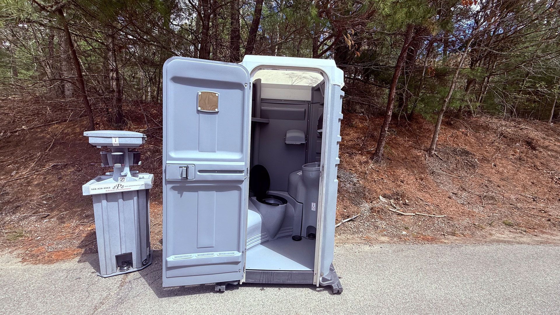 Open portable toilet beside a trash can on a wooded roadside