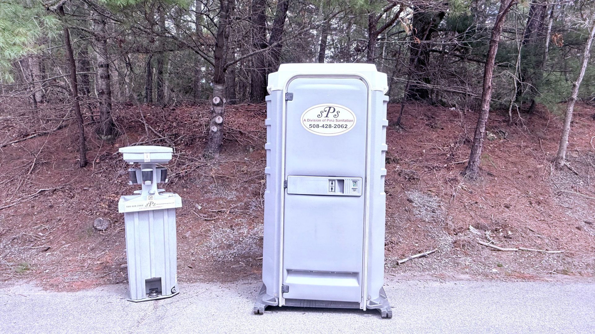 Two portable toilets beside a handwashing station on a roadside, with woods in the background.