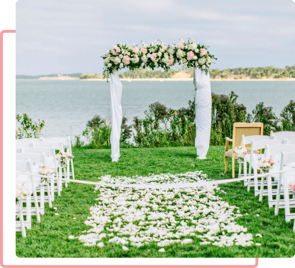 Outdoor wedding ceremony setup with white chairs, a floral archway, and a petal-strewn aisle overlooking a large lake.