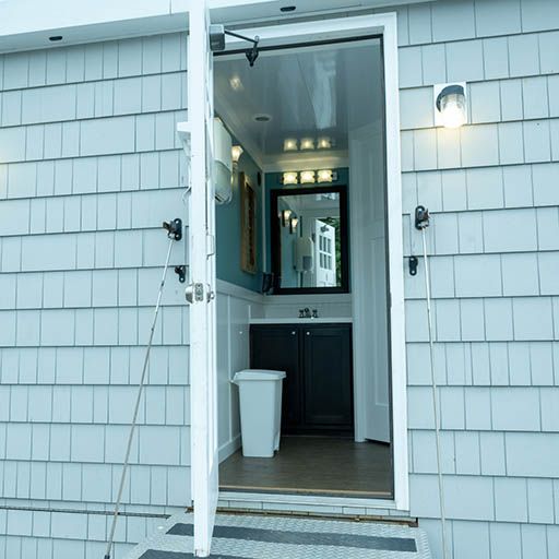 Open front door of a gray house revealing a lit hallway and white trash bin inside.