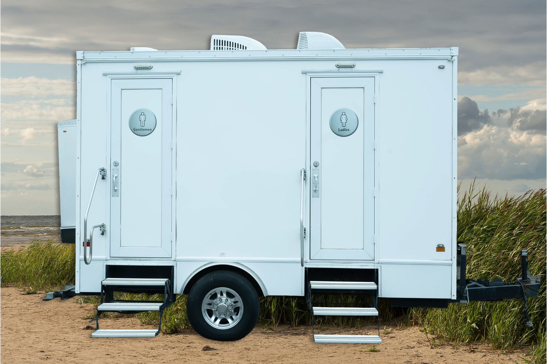 White portable restroom trailer with two doors and stairs, parked on gravel under a cloudy sky.