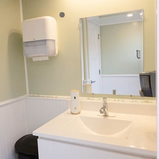 Bright restroom with white sink, mirror, soap dispenser, and black trash can beneath a paper towel dispenser
