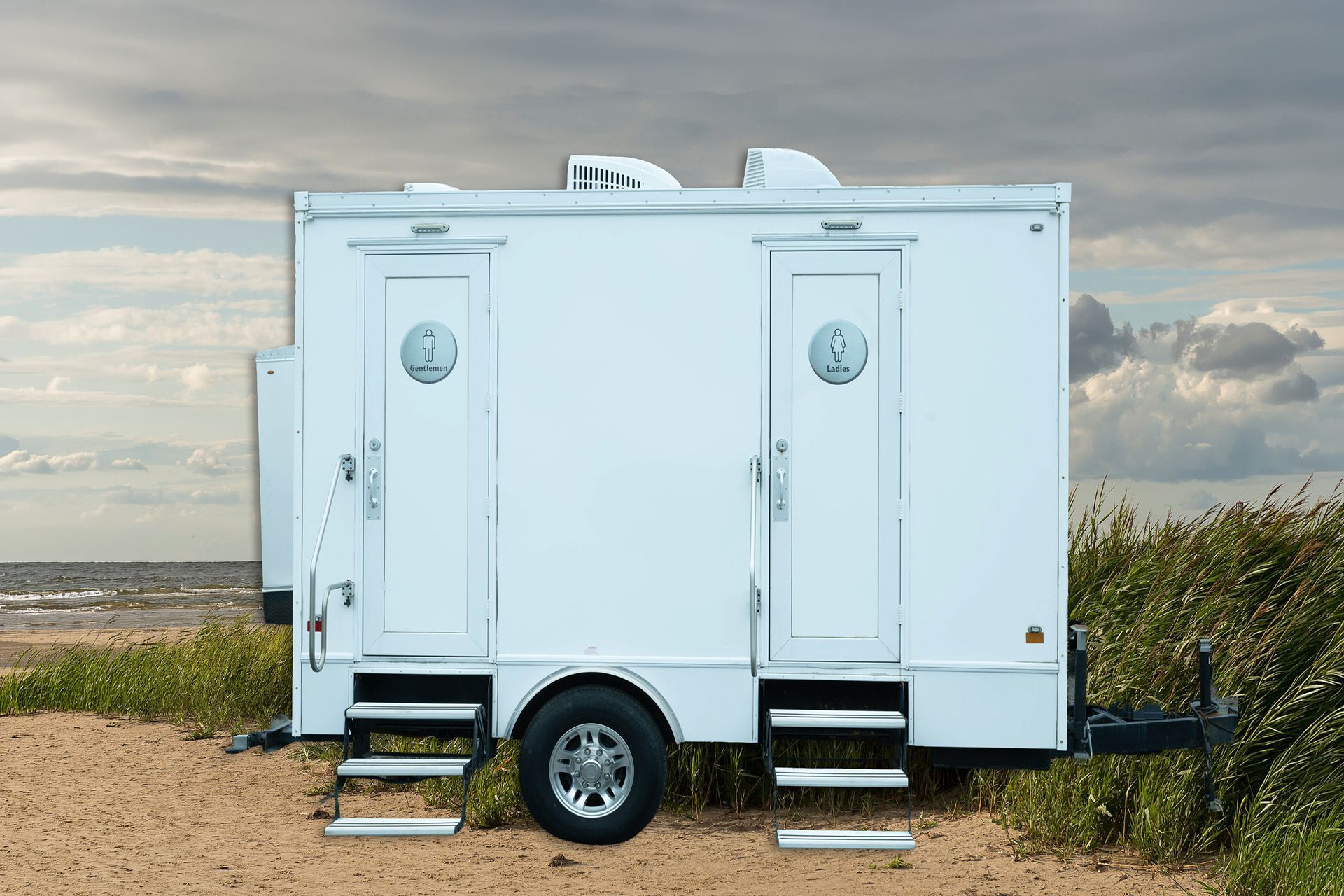 White mobile restroom trailer parked on a sandy beach with a cloudy sky backdrop.