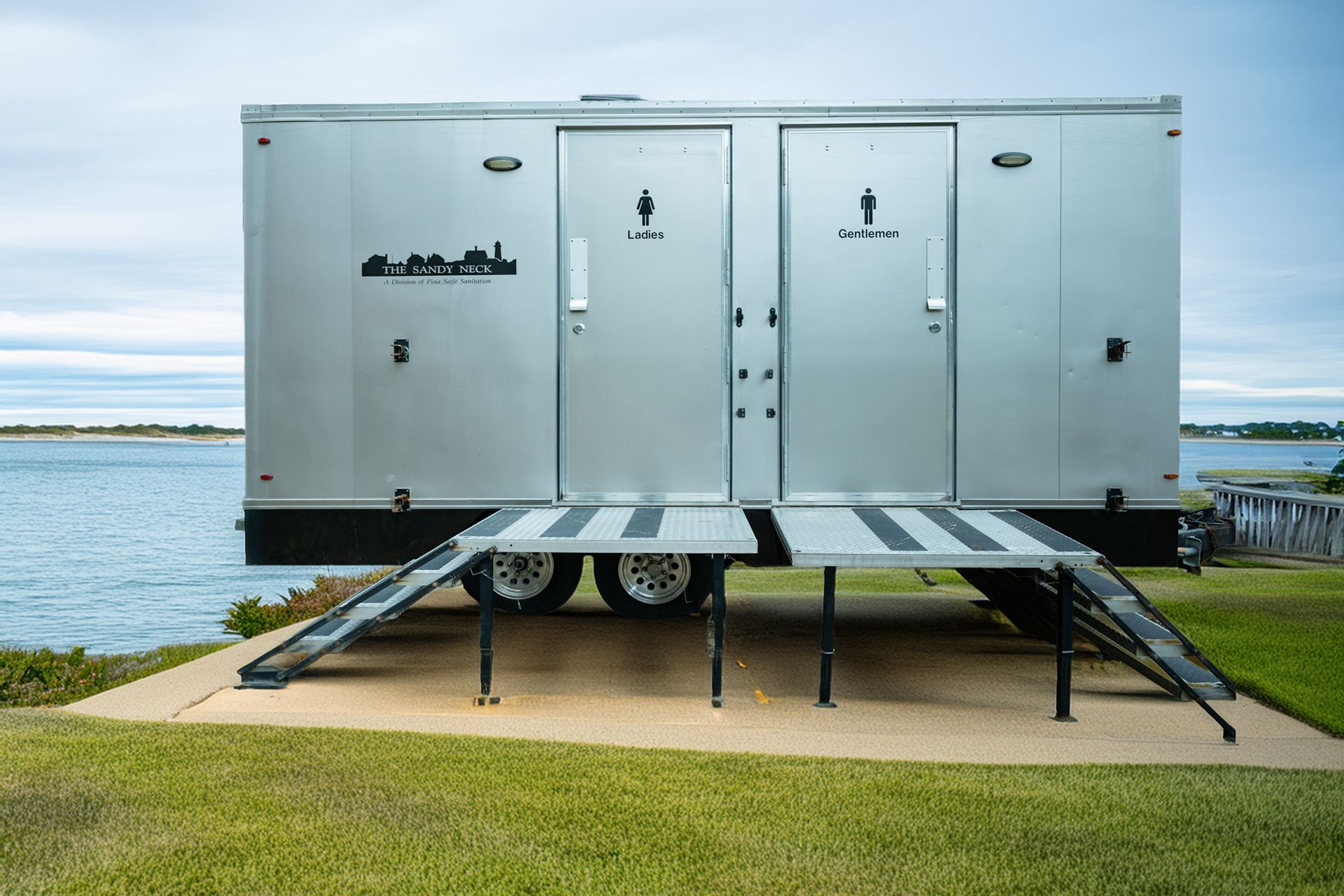 Silver portable restroom trailer with ramps beside a body of water and grassy shore.