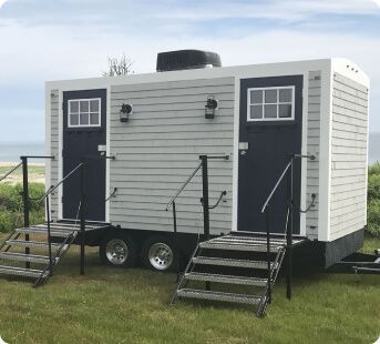 Portable restroom trailer with light gray shingled siding, two black doors with windows, and metal entry stairs.