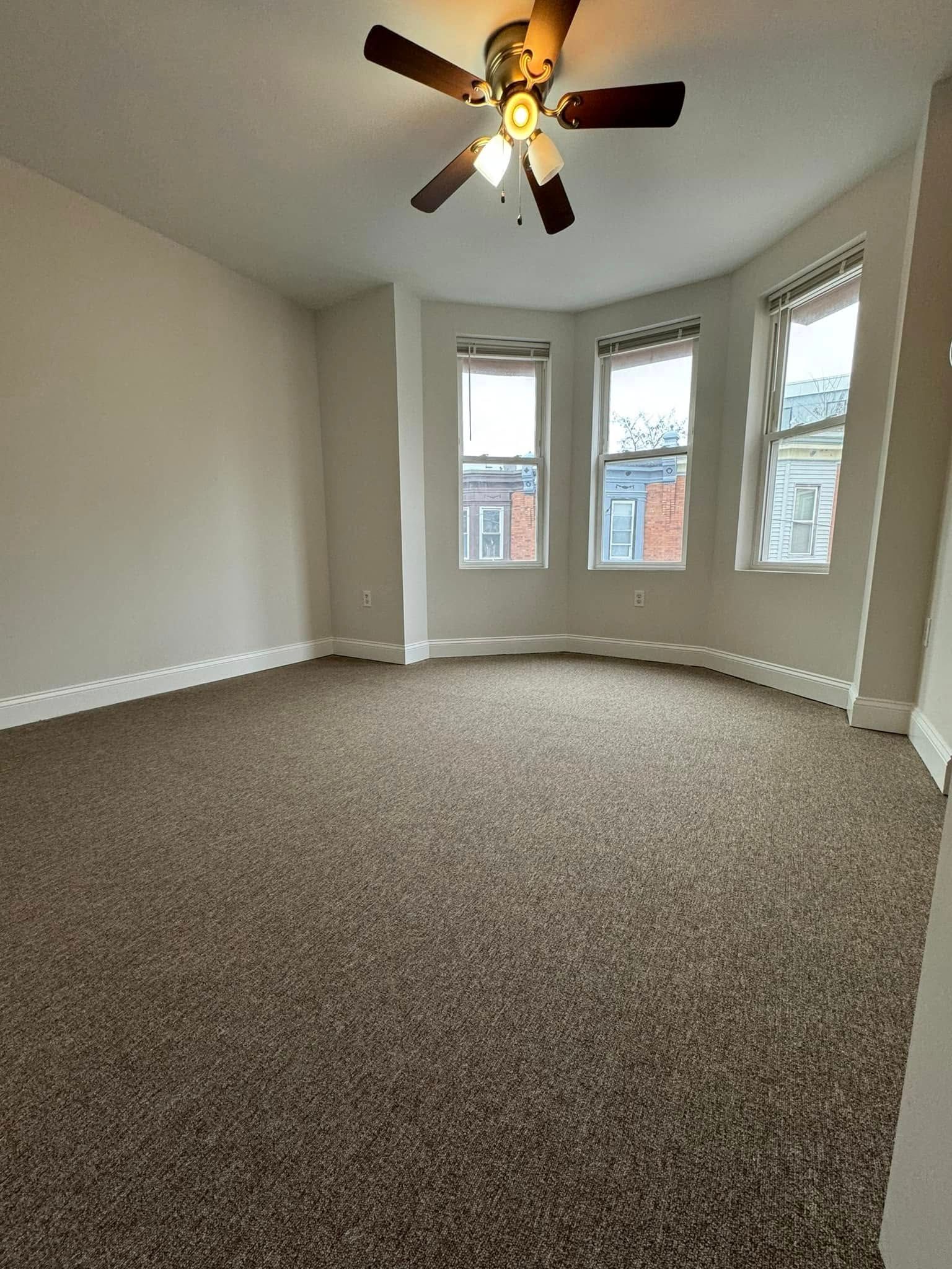 Empty carpeted bedroom with bay windows, white walls, and a ceiling fan.