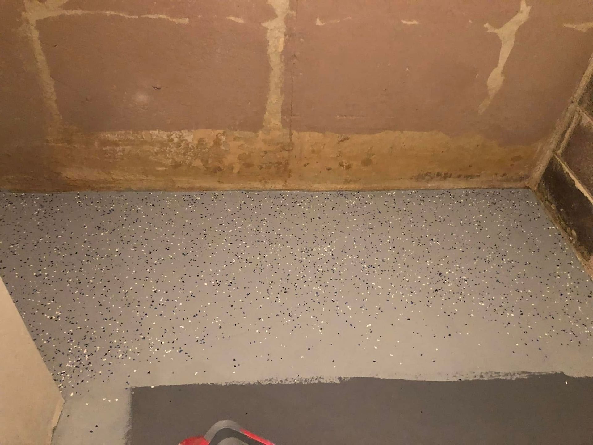 Small speckled floor and tan wall corner, with a dark mat and red-handled tool at the bottom.