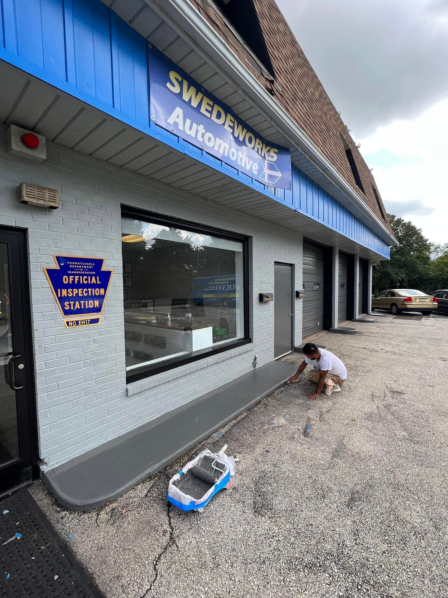 Gray auto body shop exterior with a person crouching by the wall on a gravel lot.