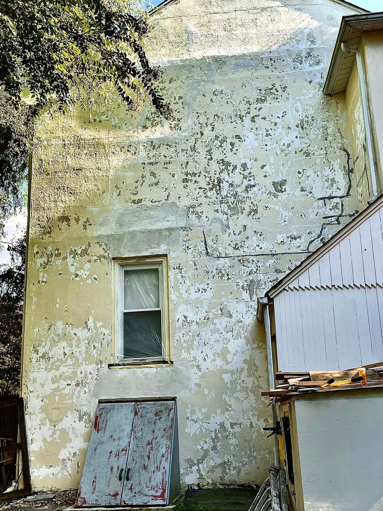 Weathered white stucco building facade with a small window and boarded lower door beside a tree.