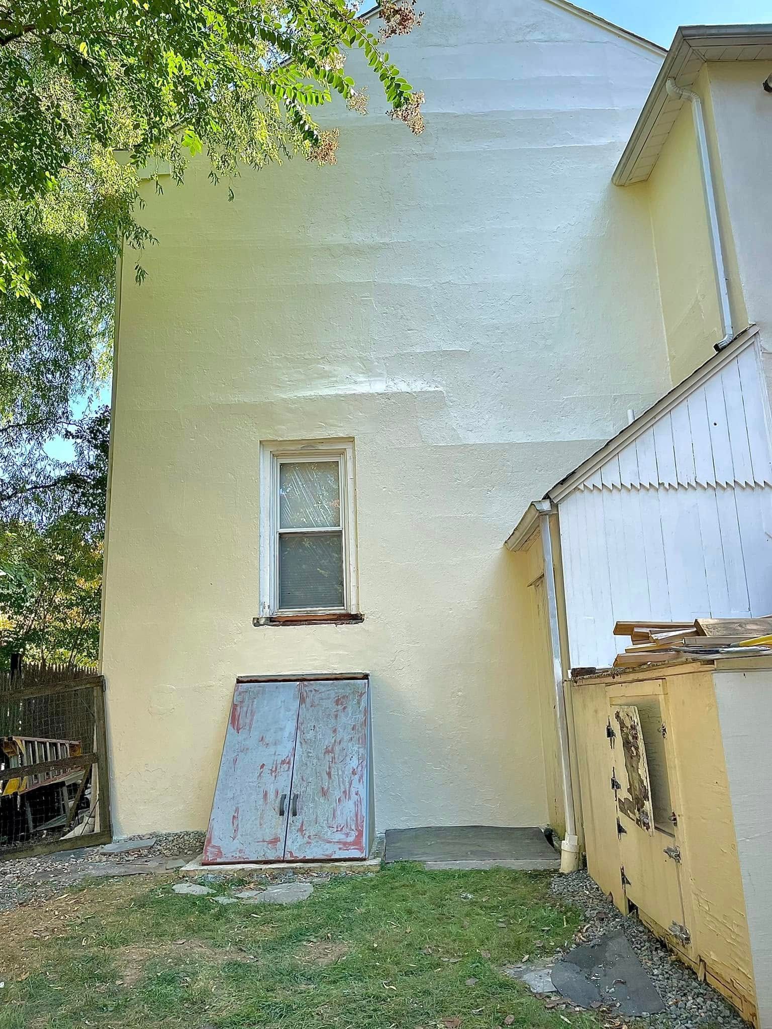 Narrow backyard view of a pale yellow house wall, small window, grass, and a leaning metal sheet below the window