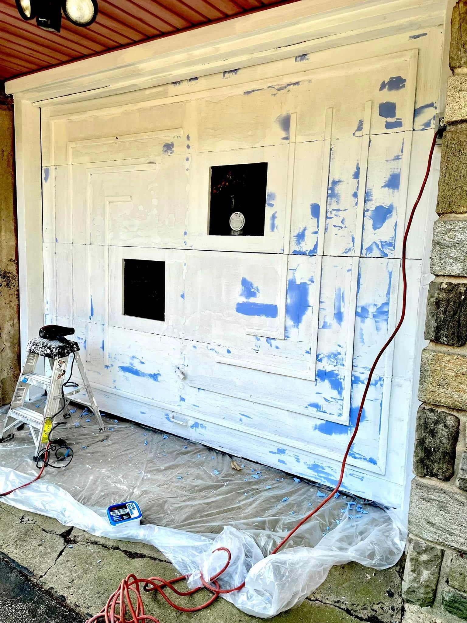 Painted garage wall with two black square openings, masking plastic, and a stepladder during prep work
