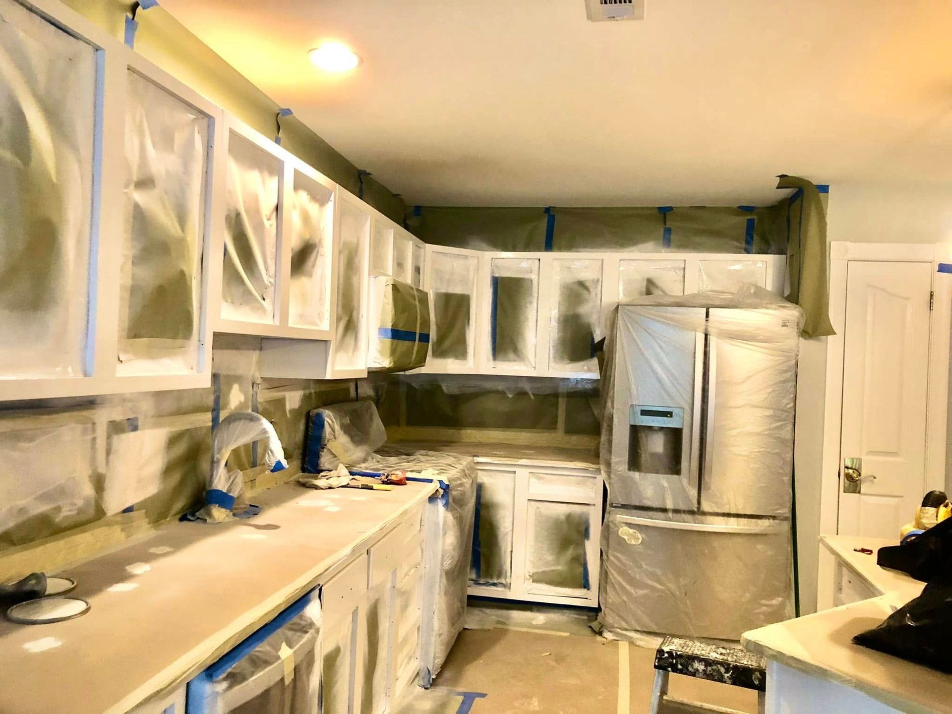 Kitchen under renovation with cabinets and appliances covered in plastic sheeting and painter’s tape.