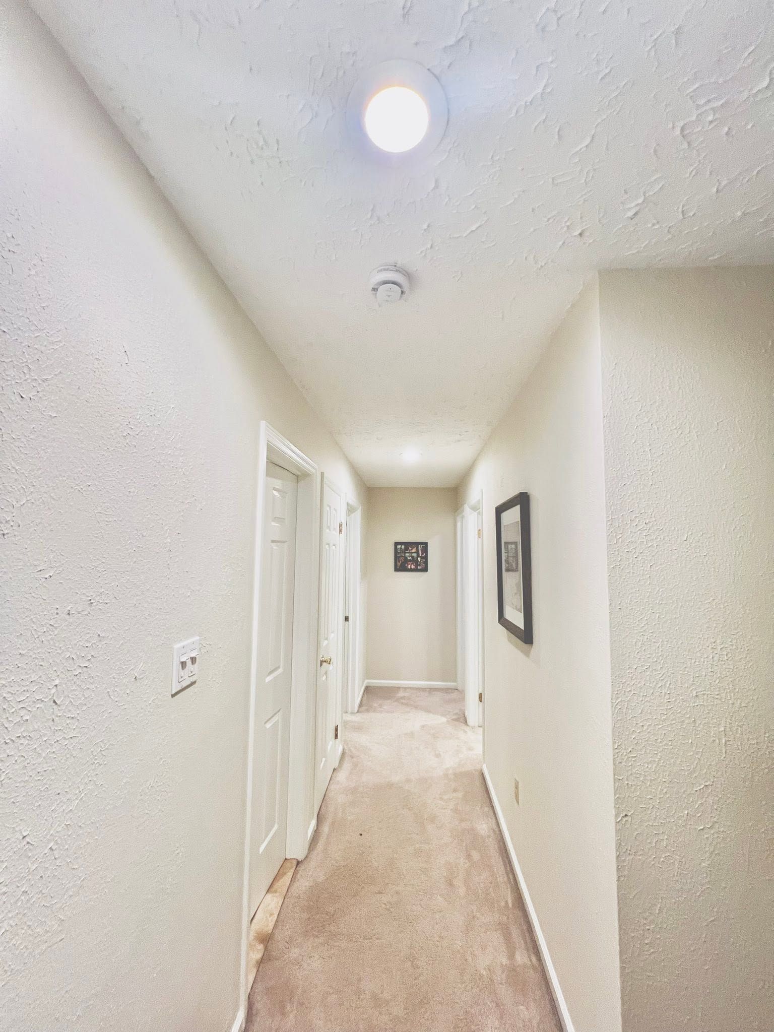 Narrow white hallway with textured walls, beige carpet, ceiling lights, and framed wall art