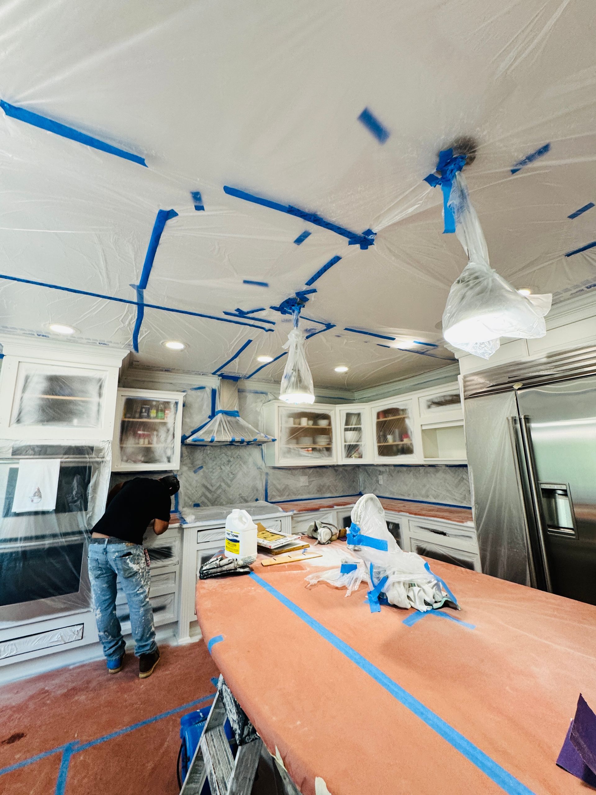 Kitchen under renovation with blue painter’s tape, covered counters, and a worker standing near the island
