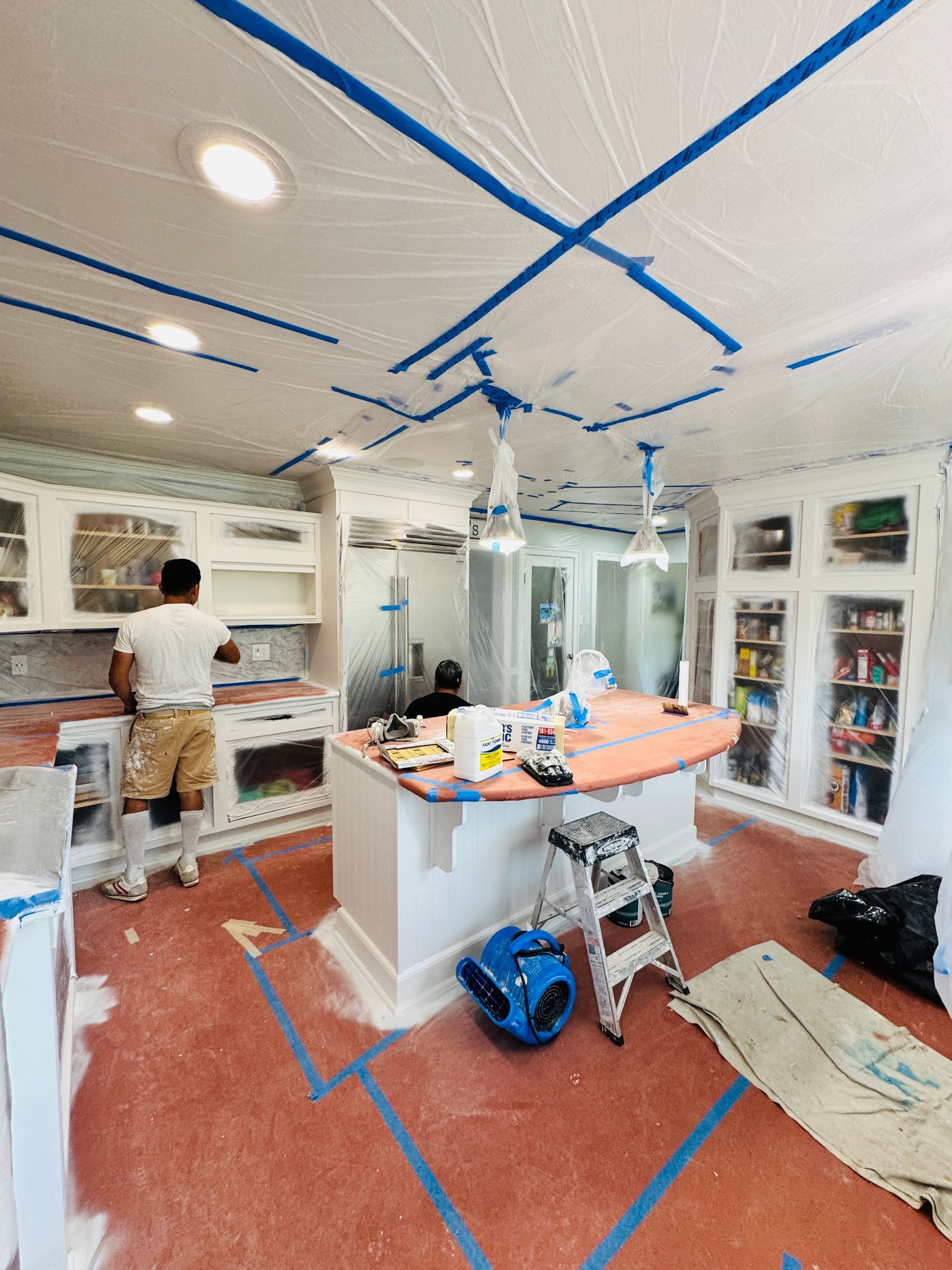 Bright kitchen under renovation, with blue painter’s tape on walls and ceiling and a person working near the sink.