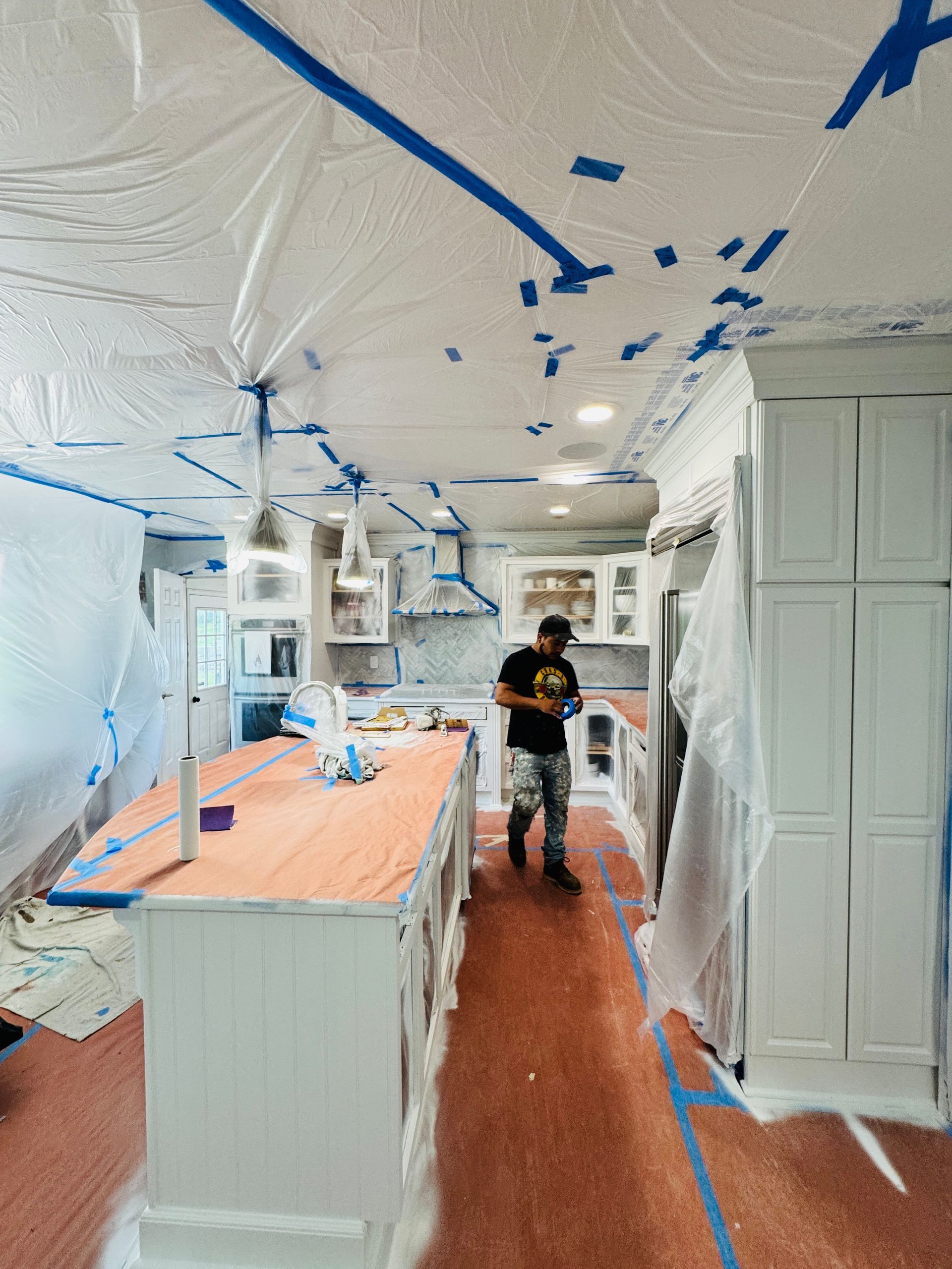 Kitchen under renovation, with blue tape, plastic-covered cabinets, and a worker standing in the center.