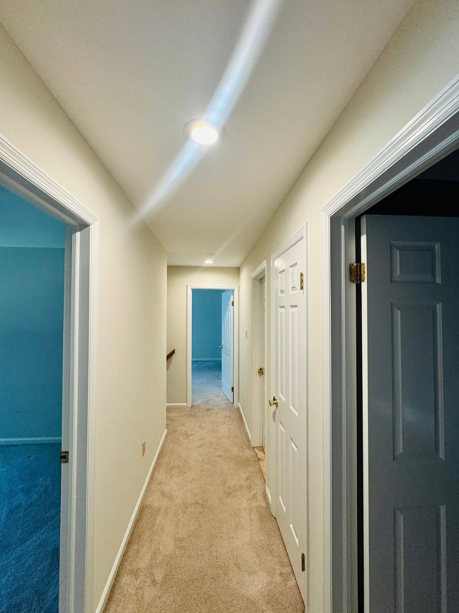 Narrow carpeted hallway with white walls and multiple open doorways, including a blue room at the end.