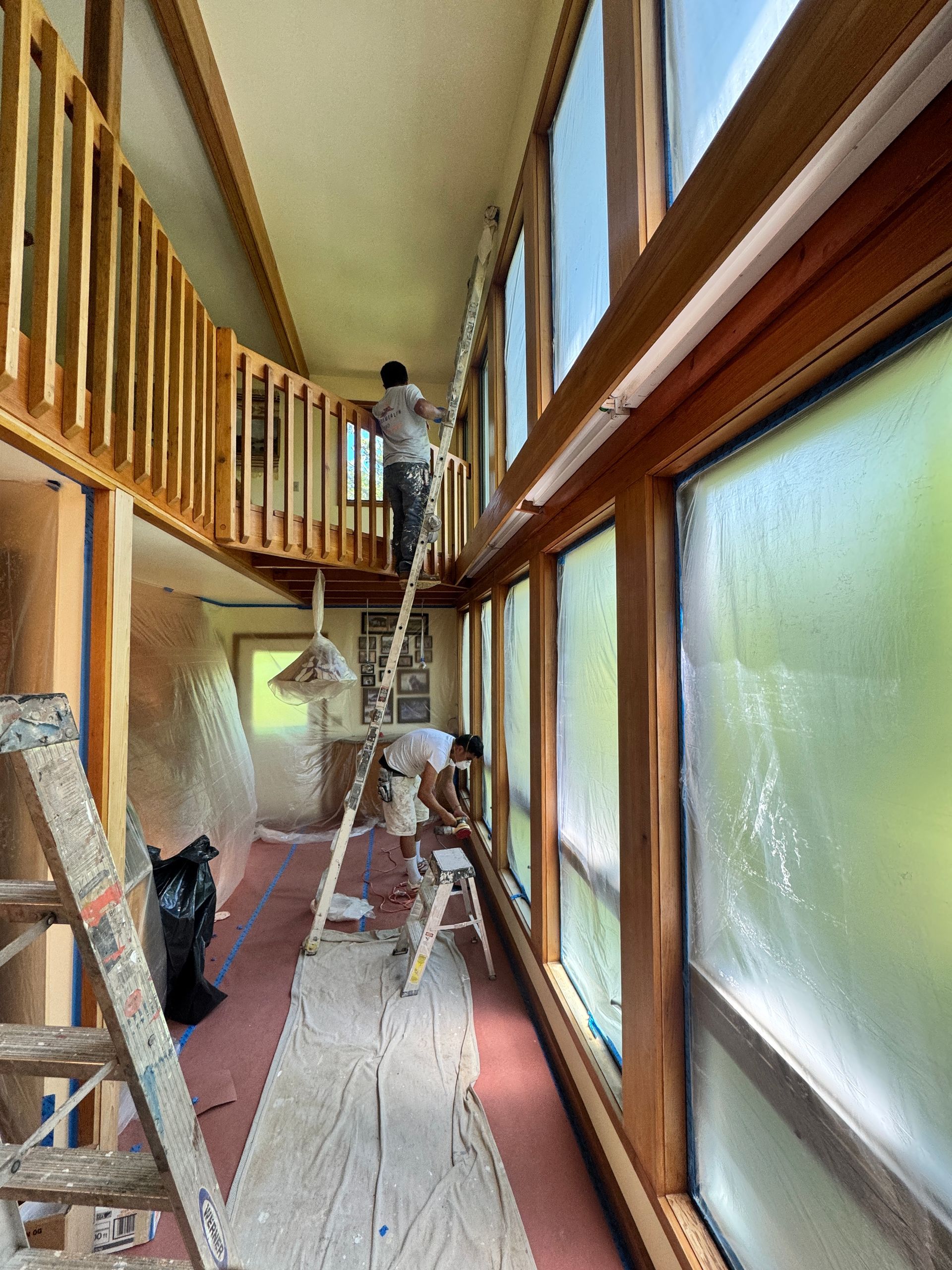 Worker on a ladder in a narrow hallway with wood trim and frosted windows, doing interior renovation work