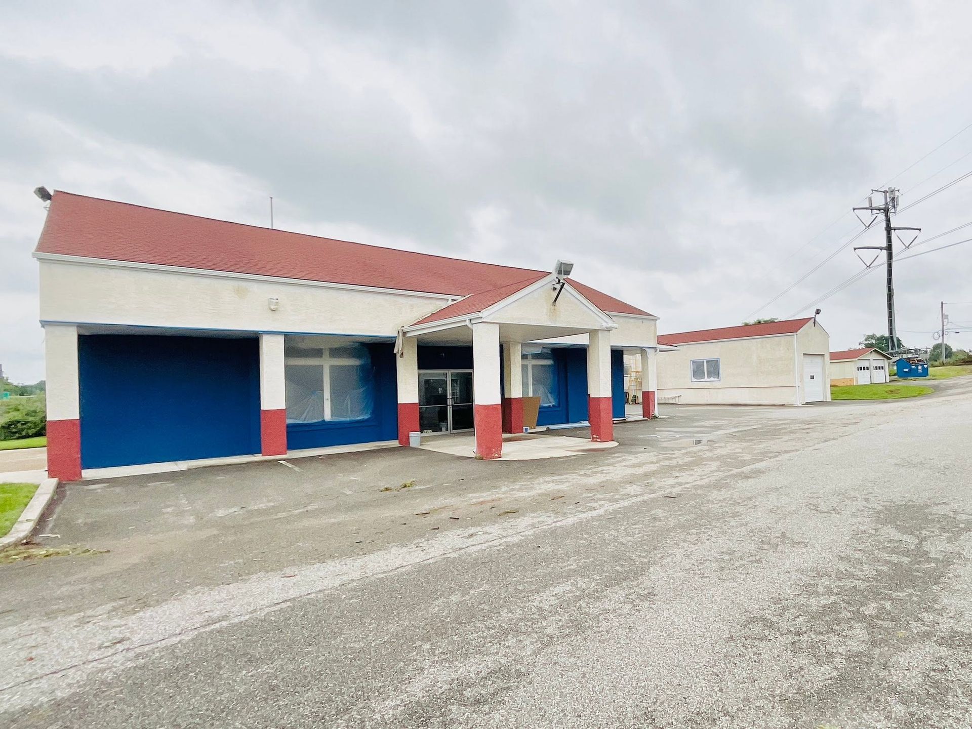 Small roadside building with red roof, blue garage doors, and a gravel lot under cloudy skies