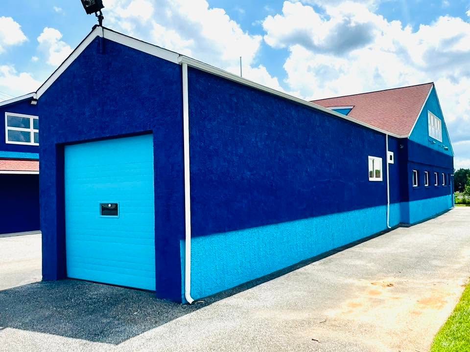 Bright blue industrial building with a large garage door and long side wall under a partly cloudy sky