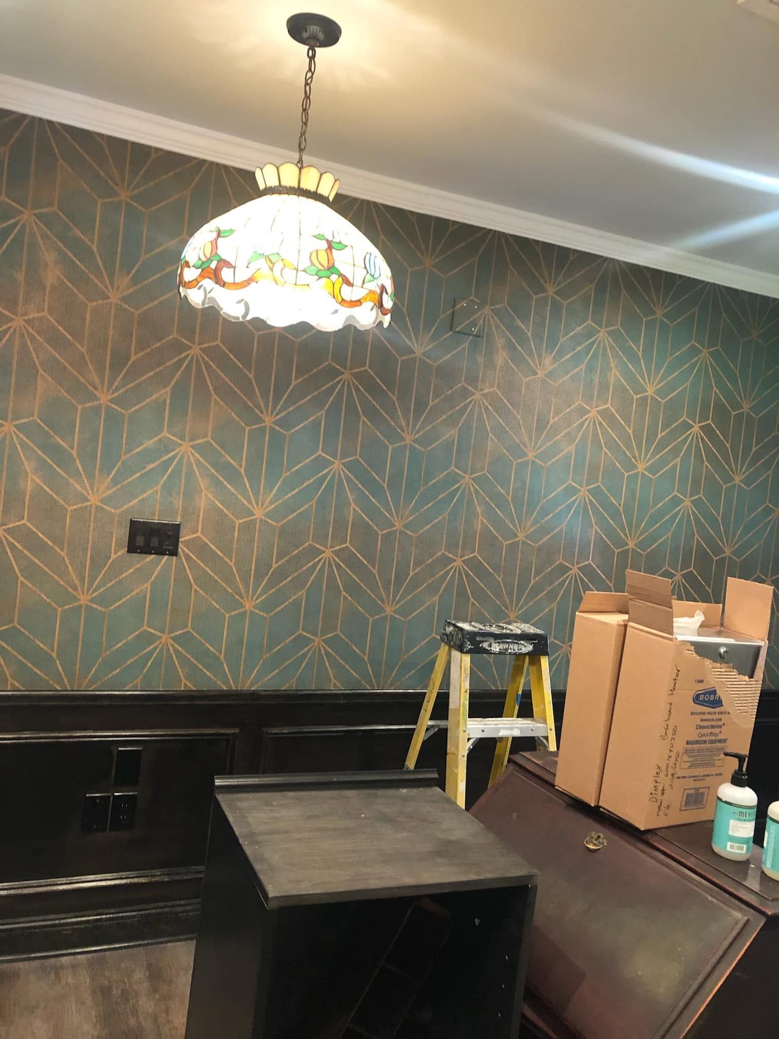 Dim room with patterned wallpaper, hanging stained-glass lamp, workbench, ladder, and stacked boxes under renovation.