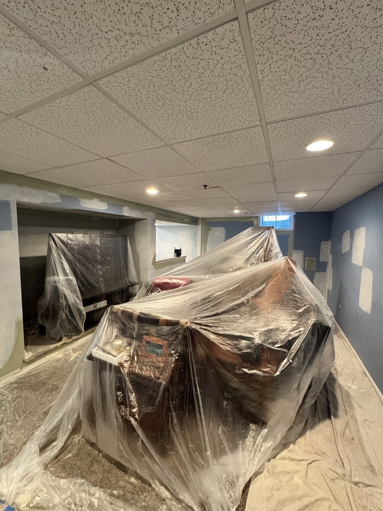 Furniture wrapped in plastic in a room with a drop ceiling and blue walls