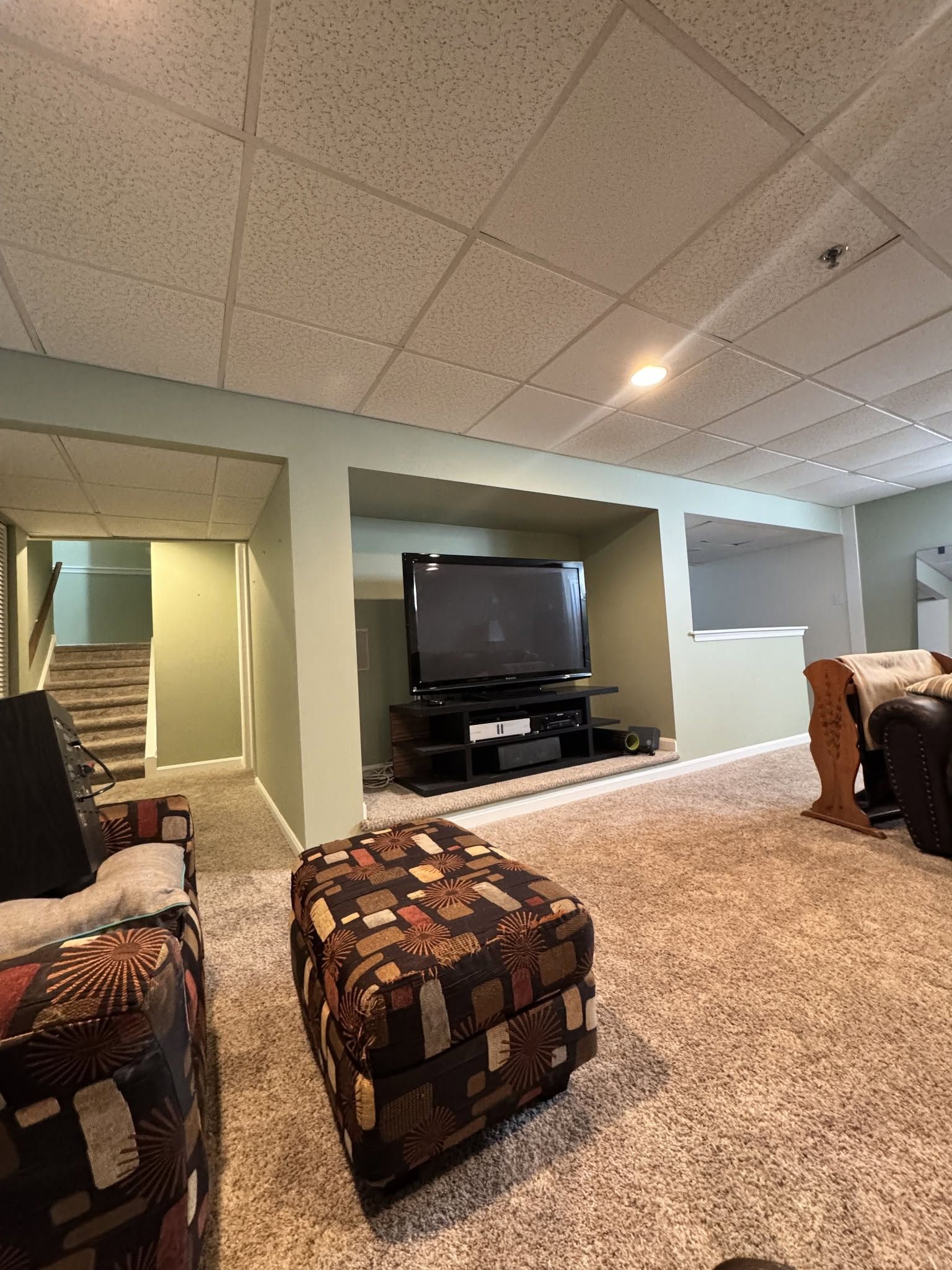 Basement family room with carpet, patterned ottoman, TV, and staircase leading up
