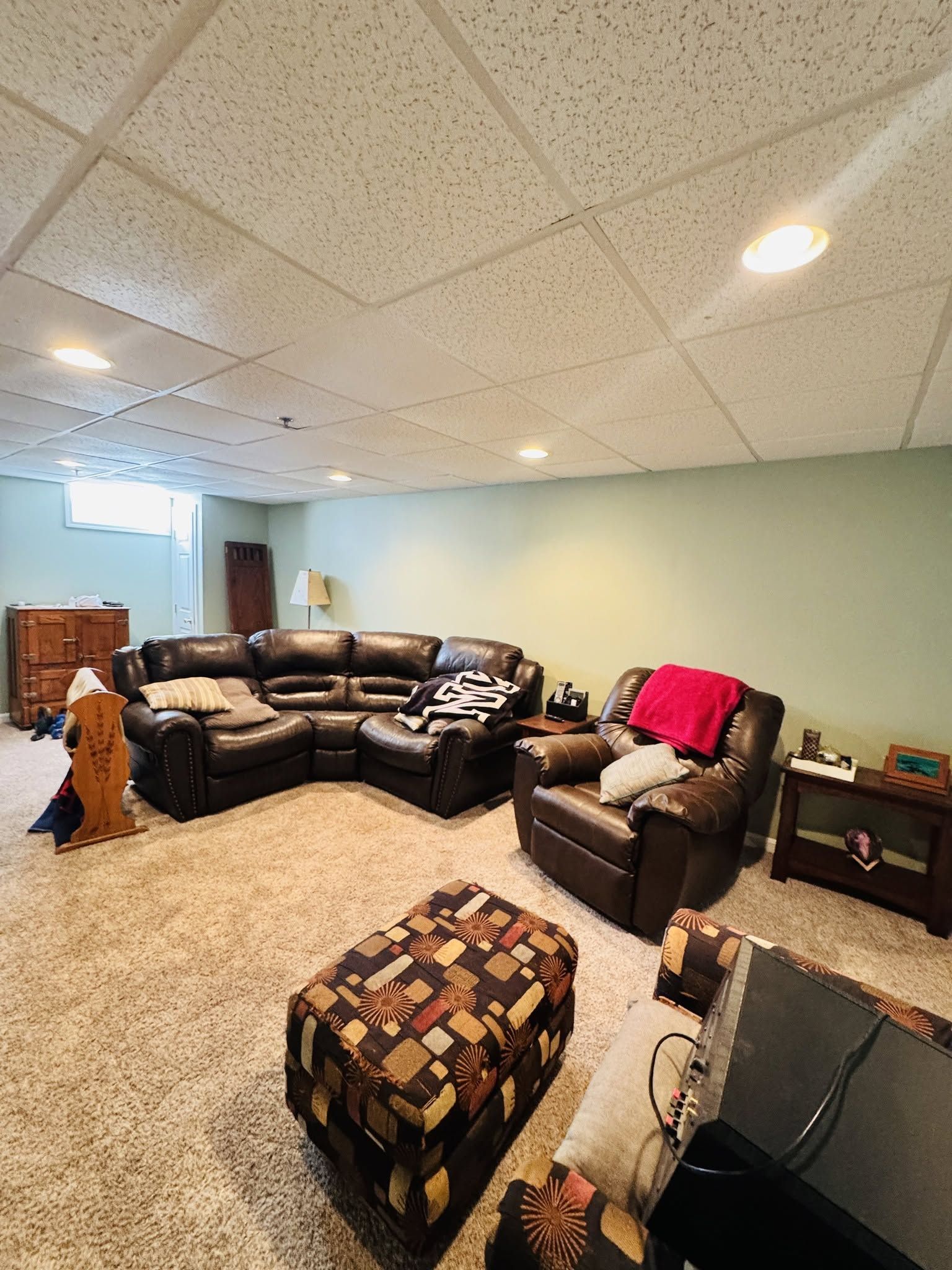 Carpeted living room with dark leather sectional, recliners, ottoman, TV, and pale green walls.