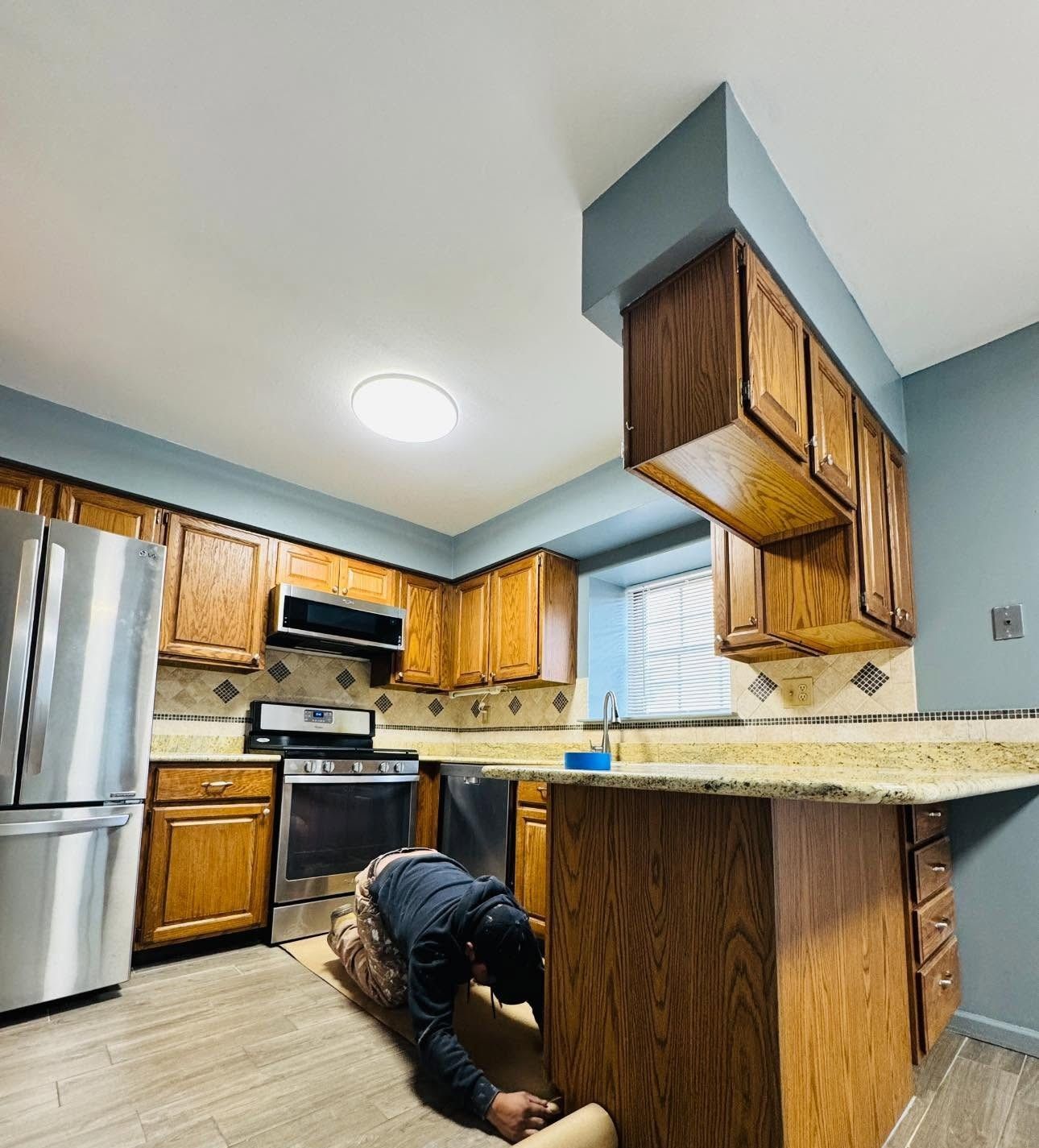 Person crouched under a kitchen island in a modern kitchen with wood cabinets and stainless steel appliances