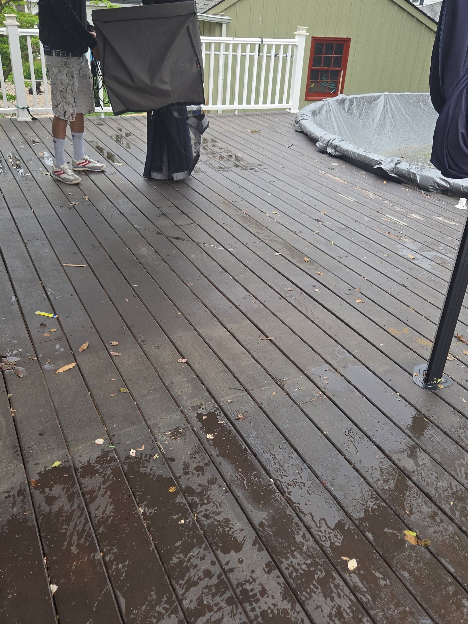 Rainy deck with two people standing near a white railing and a covered grill, wet wooden floor in foreground