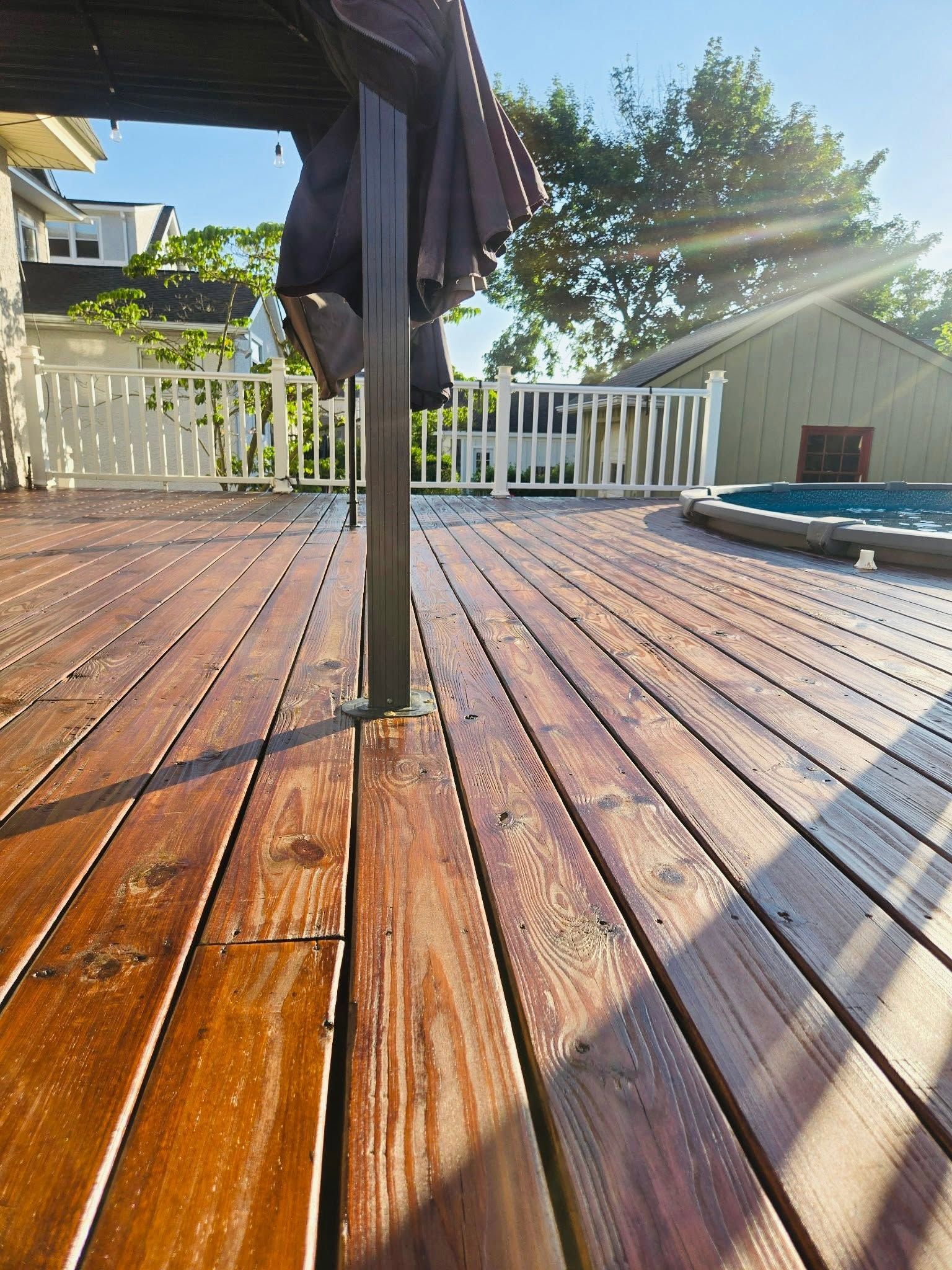 Sunlit wooden deck with white railing, patio umbrella, and swimming pool in the background