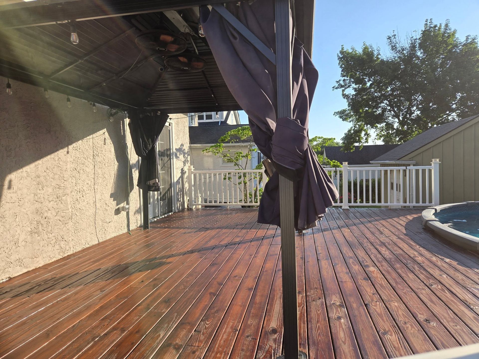 Wooden patio deck under a covered pergola with a closed dark umbrella and white fence in sunlight