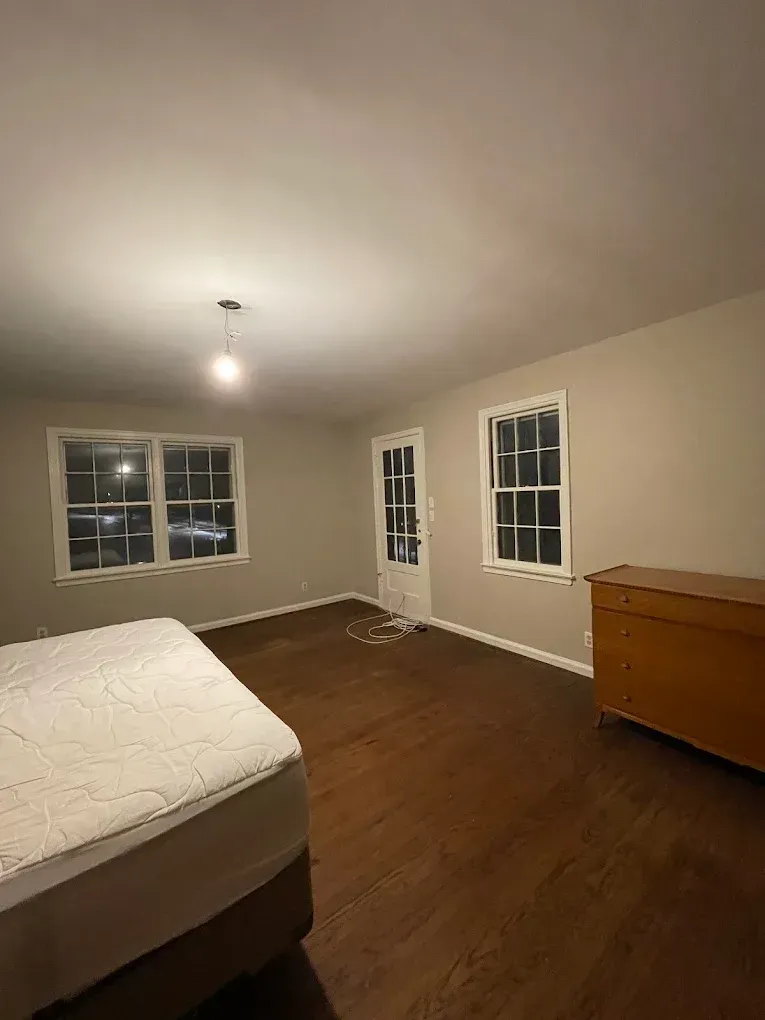 Empty bedroom with wood floors, beige walls, two windows, a door, and a dresser.