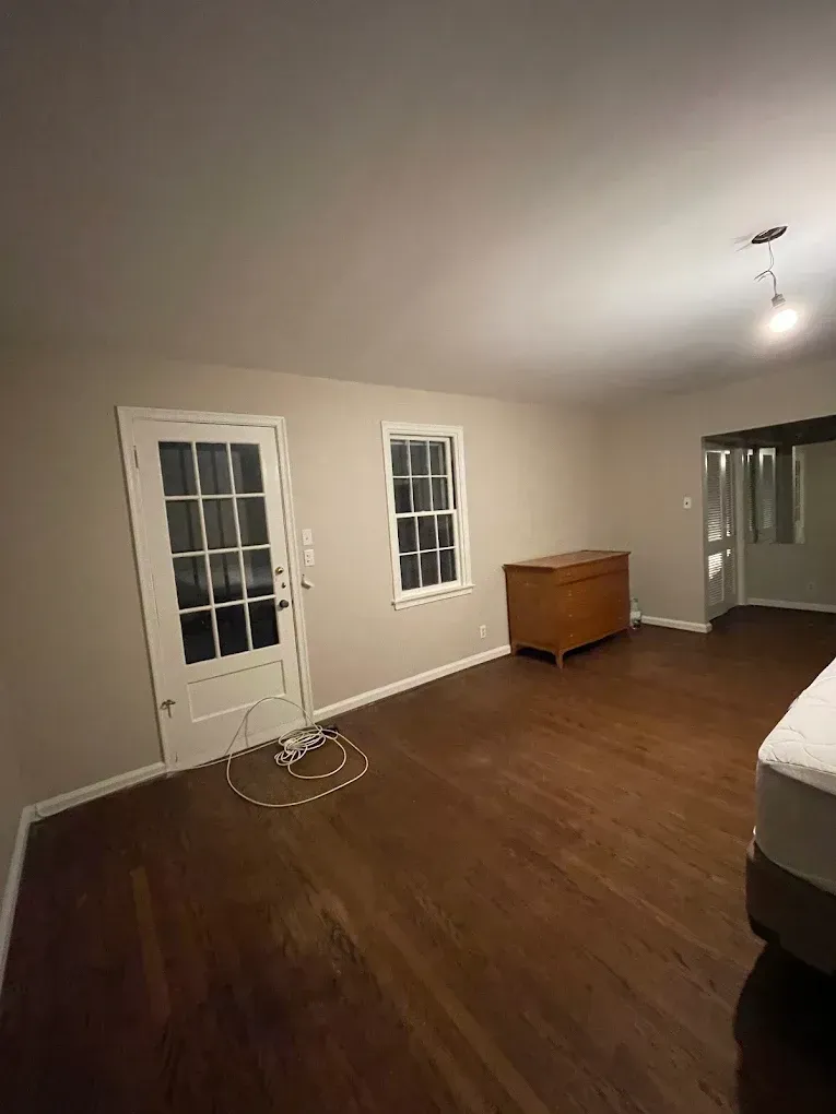 Empty living room with hardwood floor, white walls, French doors, window, and a wooden dresser.