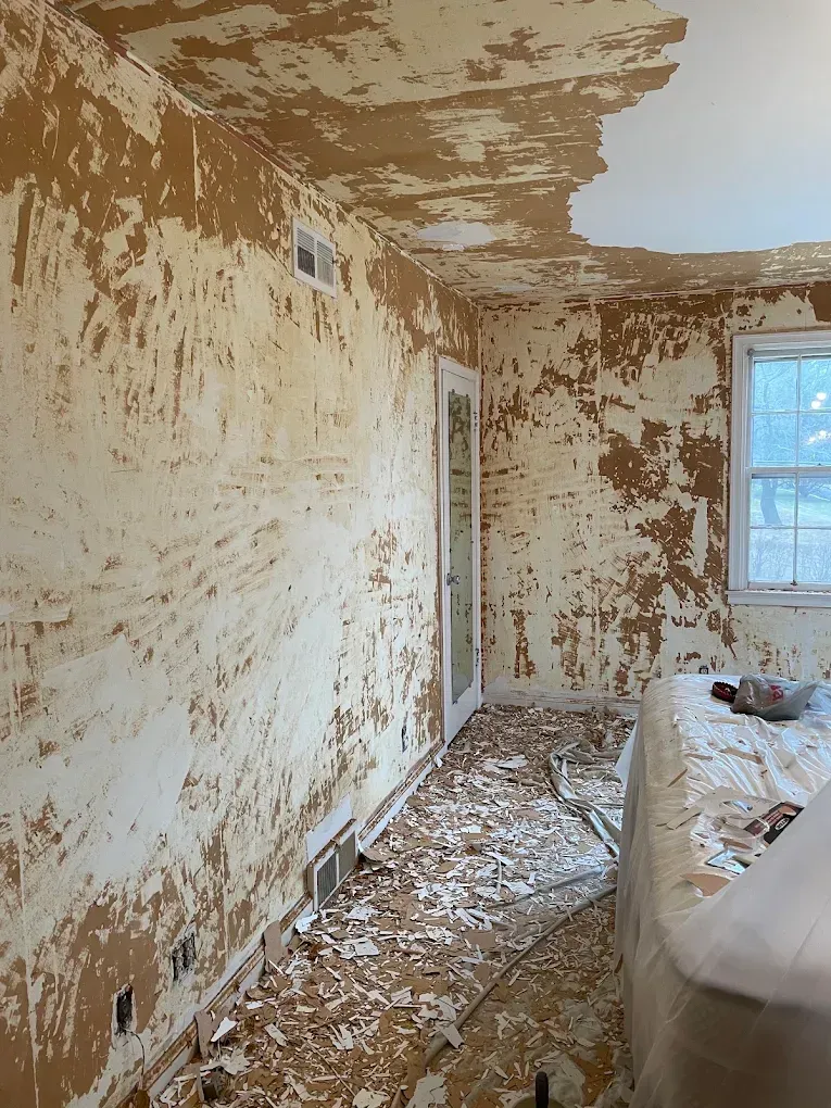 A room under renovation with peeling plaster, debris on the floor, and a window letting in light.