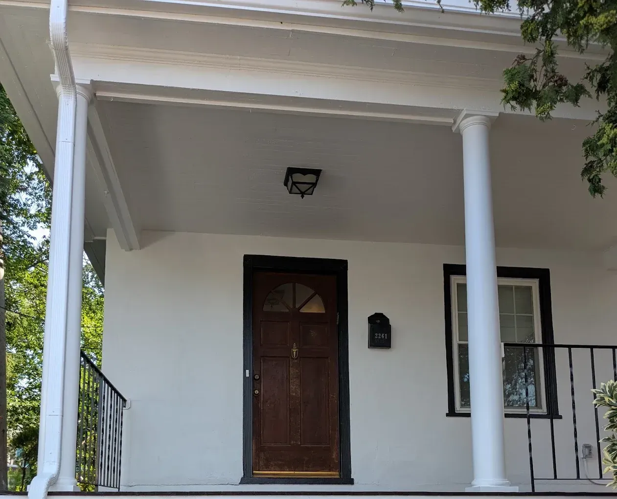 Front porch of a white house with a brown door, two columns, and black railings