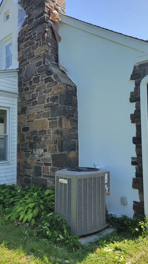 Stone chimney on a white house exterior beside a wooden air-conditioning unit, with grass and shrubs below.
