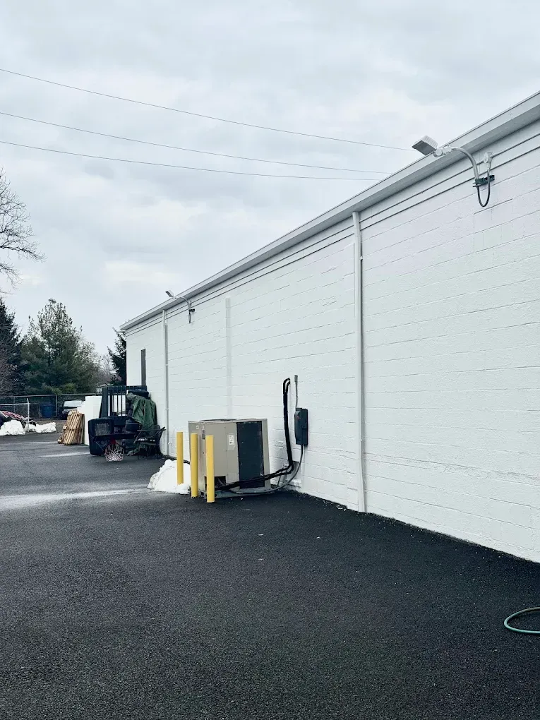 Side of a white industrial building with a forklift and yellow equipment on gravel.