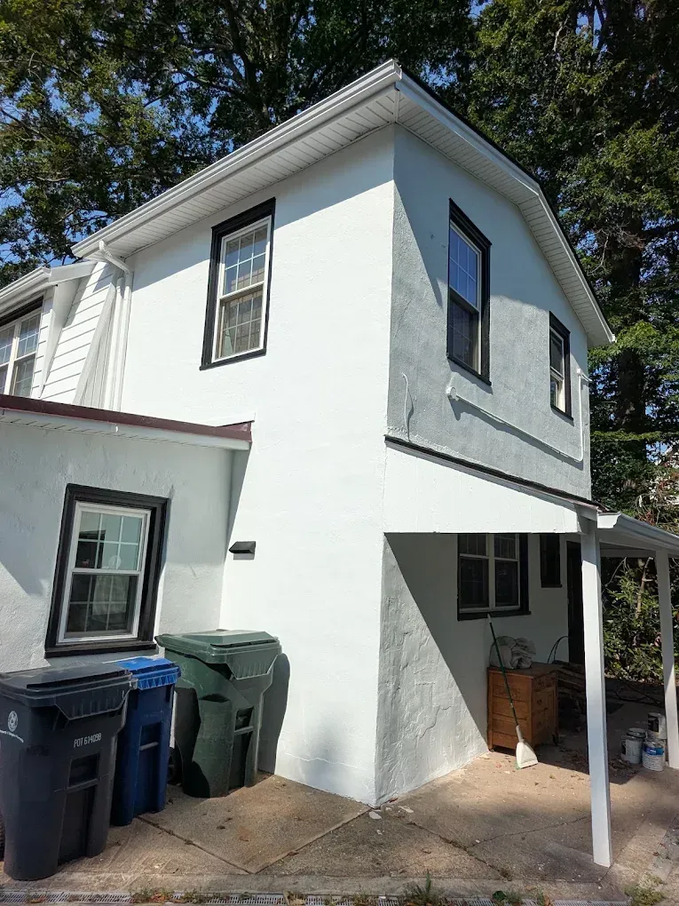 White two-story house exterior with black-trim windows, side porch, and trash bins by the driveway