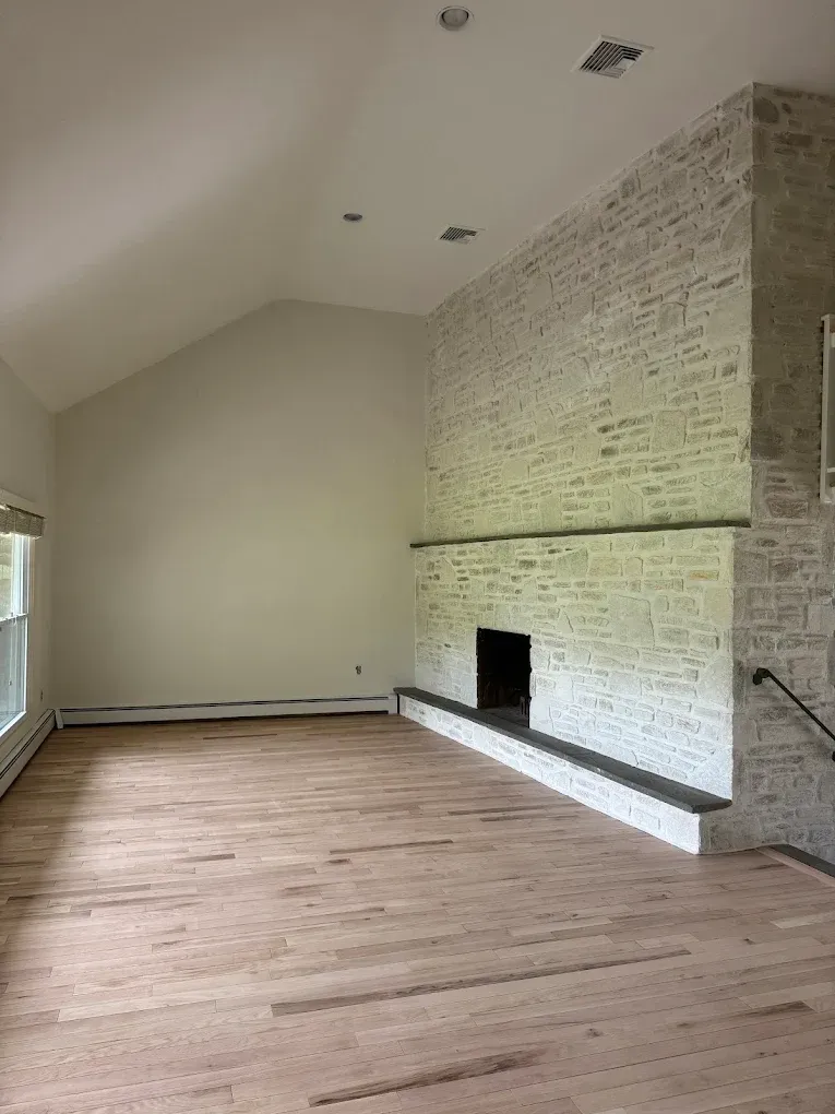 Empty living room with hardwood floors, vaulted ceiling, and a white brick fireplace on the right.